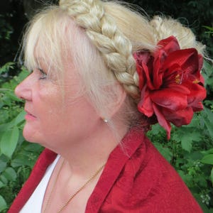 May include: A woman with blonde hair styled in a braid crown and a bun, wearing a red shawl and a gold necklace. A large red flower is pinned to her hair.