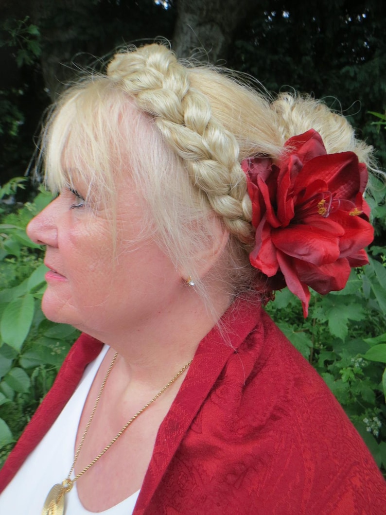 May include: A woman with blonde hair styled in a braid crown and a bun, wearing a red shawl and a gold necklace. A large red flower is pinned to her hair.