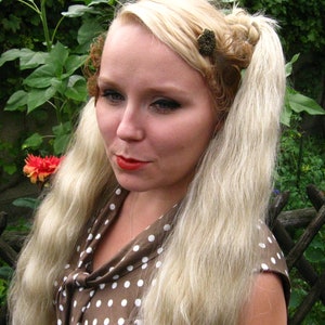 May include: A woman with long blonde hair styled in two braids with decorative hair clips. She is wearing a brown and white polka dot dress with a bow at the neck.