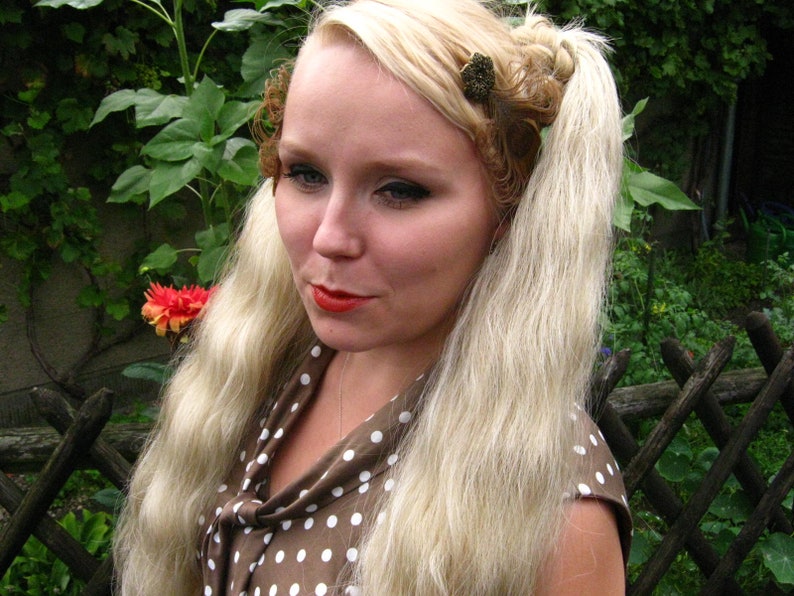 May include: A woman with long blonde hair styled in two braids with decorative hair clips. She is wearing a brown and white polka dot dress with a bow at the neck.