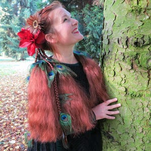 May include: A woman with long, auburn hair adorned with a red flower and peacock feathers. She is wearing a black top and a fur-like jacket with peacock feathers.