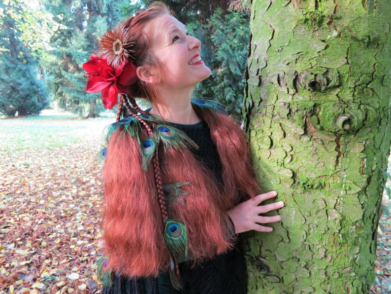 May include: A woman with long, auburn hair adorned with a red flower and peacock feathers. She is wearing a black top and a fur-like jacket with peacock feathers.