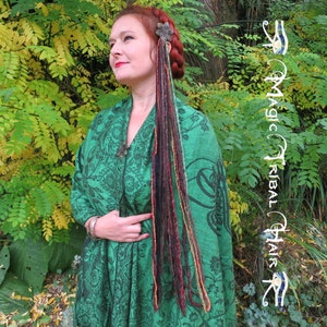 May include: A woman with long, colorful dreadlocks wearing a green shawl with a floral pattern. She is standing in front of a green bush with yellow leaves.