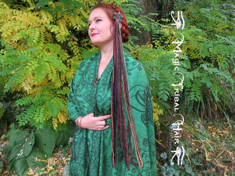 May include: A woman with long, colorful dreadlocks wearing a green shawl with a floral pattern. She is standing in front of a green bush with yellow leaves.