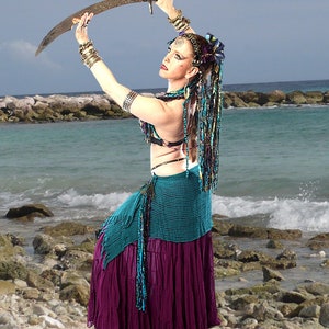 May include: A woman in a turquoise and purple skirt is holding a scimitar sword in front of a beach scene. She is wearing a turquoise bra and a beaded necklace. Her hair is long and braided with colorful beads.