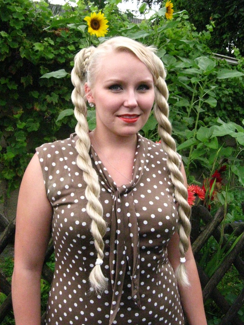 May include: A woman with blonde hair styled in two braids wears a brown dress with white polka dots. The dress has a bow at the neckline.