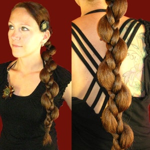 Op de afbeelding: Een vrouw met lang bruin haar dat is gestyled in een gevlochten paardenstaart met grote, ronde bulten. Het haar is van haar gezicht weggetrokken en vastgezet met een zwart elastiek. Ze draagt een zwart tanktop met een kruisband-ontwerp.