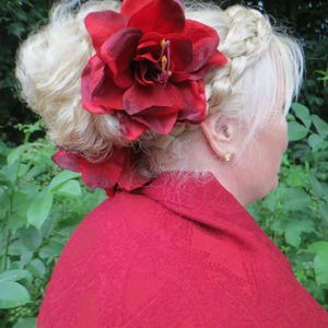 May include: A woman with blonde hair styled in a bun with a braid, wearing a red shawl and a large red flower hair accessory.