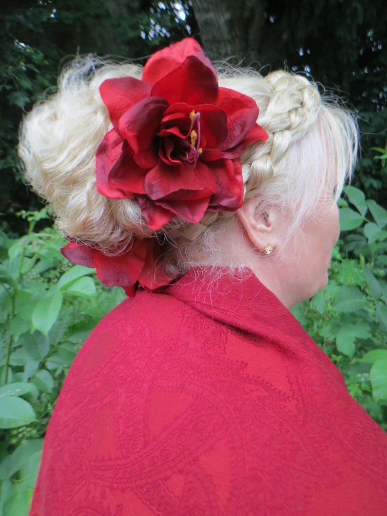 May include: A woman with blonde hair styled in a bun with a braid, wearing a red shawl and a large red flower hair accessory.