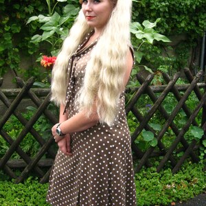 May include: A woman with long blonde hair wearing a brown and white polka dot dress. She is standing in front of a wooden fence with green plants behind her.