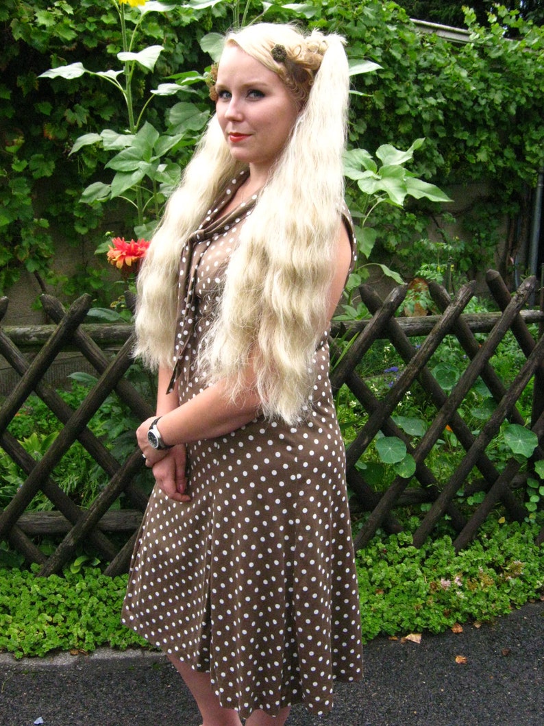 May include: A woman with long blonde hair wearing a brown and white polka dot dress. She is standing in front of a wooden fence with green plants behind her.