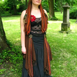 May include: A woman with long, red and brown dreadlocks wears a black lace top with a red flower, a brown vest, and a black skirt. She is standing in a grassy area with trees in the background.