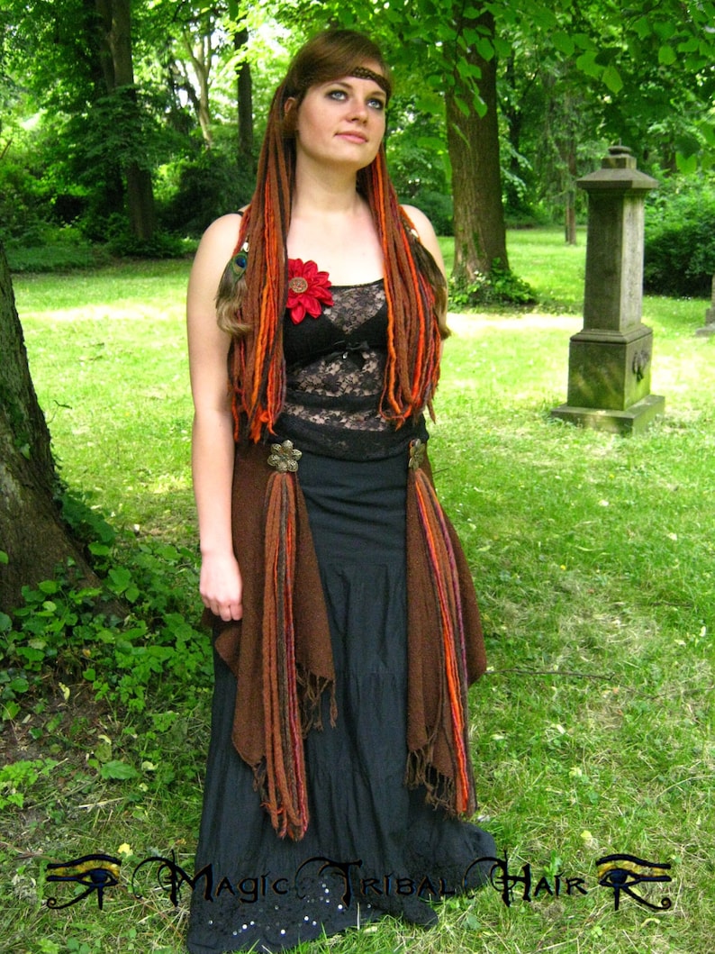 May include: A woman with long, red and brown dreadlocks wears a black lace top with a red flower, a brown vest, and a black skirt. She is standing in a grassy area with trees in the background.