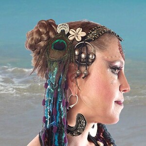 May include: A woman with brown hair styled in a braid wears a silver and brown headpiece with a peacock feather and a white flower. She is wearing large silver earrings.