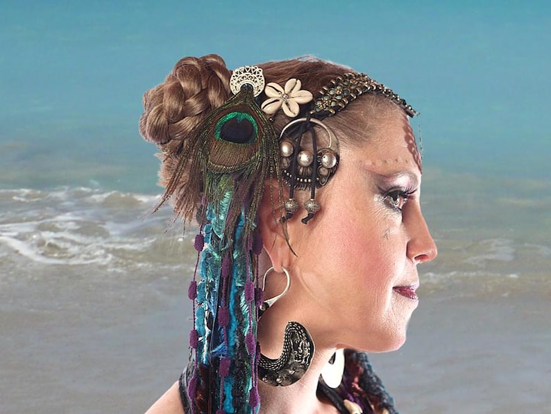 May include: A woman with brown hair styled in a braid wears a silver and brown headpiece with a peacock feather and a white flower. She is wearing large silver earrings.