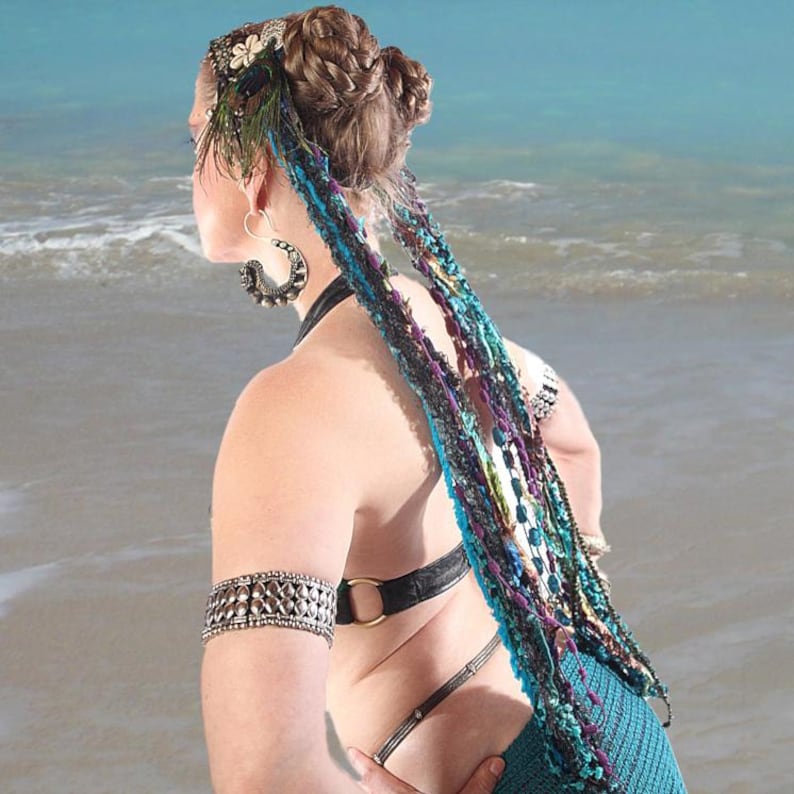 May include: A woman with long, braided hair adorned with a beaded headband and feathers. She is wearing a turquoise crochet skirt and a black leather bra with a silver metal arm cuff. The background is a blurred image of a beach.