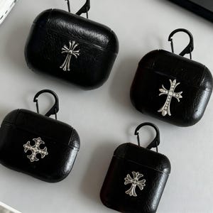 May include: Four black AirPod cases, each with a silver cross design. The cases have a textured finish and a black carabiner clip. The crosses are in different styles, some with a studded appearance.