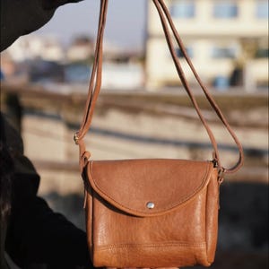 Puede incluir: Un bolso bandolera de cuero marrón con cierre de solapa y correa ajustable. El bolso tiene forma rectangular con una solapa redondeada asegurada por un botón plateado. La correa es fina y marrón.