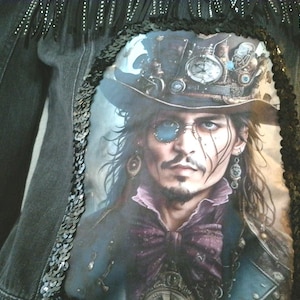 May include: Black denim jacket with a large graphic print of a man wearing a hat and glasses. The jacket is embellished with black sequins and fringe along the shoulders and edges. The graphic features a man with long hair, a purple bow tie, and a blue monocle.