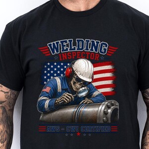 May include: A black t-shirt featuring a graphic of a skeleton wearing a hard hat and ear protection, inspecting a metal pipe with a magnifying glass. The design includes the text WELDING INSPECTOR and AWS - CWI CERTIFIED with a US flag background.