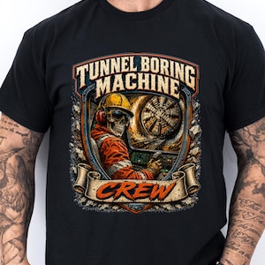 May include: A black t-shirt featuring a graphic print of a skeleton wearing a yellow hard hat, orange safety jacket, and ear protection, operating a tunnel boring machine. The text reads TUNNEL BORING MACHINE and CREW in a stylized font.