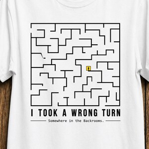 May include: A white short-sleeve t-shirt featuring a black square maze graphic with a small yellow figure inside. Below the maze, the text reads I TOOK A WRONG TURN followed by the smaller text Somewhere in the Backrooms, all in black lettering.