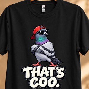 Può includere: Una t-shirt nera a maniche corte con la grafica di un piccione che indossa un berretto da baseball rosso e occhiali da sole neri. Il piccione è in piedi con le ali incrociate. Sotto l'uccello, il testo recita THAT'S COO. in grassetto bianco.