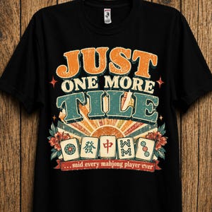 Könnte beinhalten: Ein schwarzes Kurzarm-T-Shirt mit einem Grafikdruck, der den Text "JUST ONE MORE TILE" und "...said every mahjong player ever" zeigt. Das Design enthält fünf Mahjong-Spielsteine, einen Sonnenaufgangshintergrund sowie florale Akzente in Orange, Petrol und Rot.