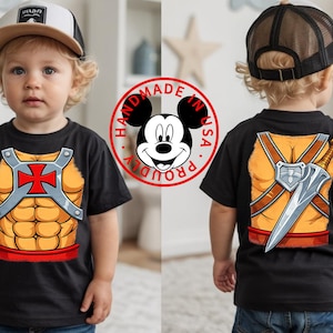May include: Black t-shirt featuring a cartoon design of a superhero's chest and back. The front displays a muscular chest with a red cross and silver accents. The back shows a sword and shield design. A "Handmade in USA" Mickey Mouse logo is visible.
