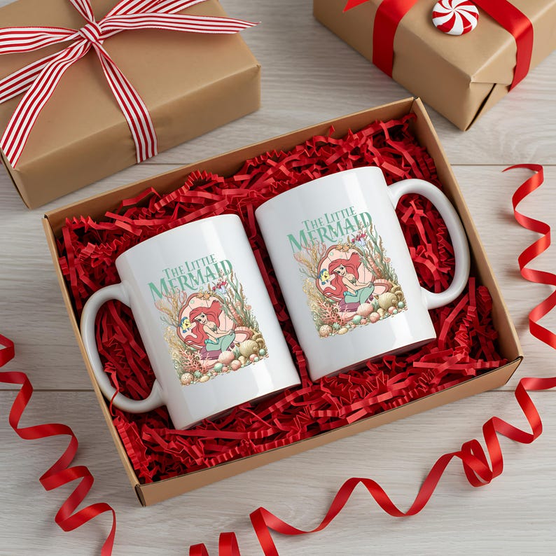 A pair of white mugs with the text "The Little Mermaid" printed on them, placed in a brown gift box filled with red shredded paper and red ribbons.
