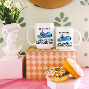 two white mugs with a cartoon character and text, placed on a pink tablecloth. There are also two doughnuts with colorful sprinkles and a white bust of a person on a pedestal. The background has a floral pattern.