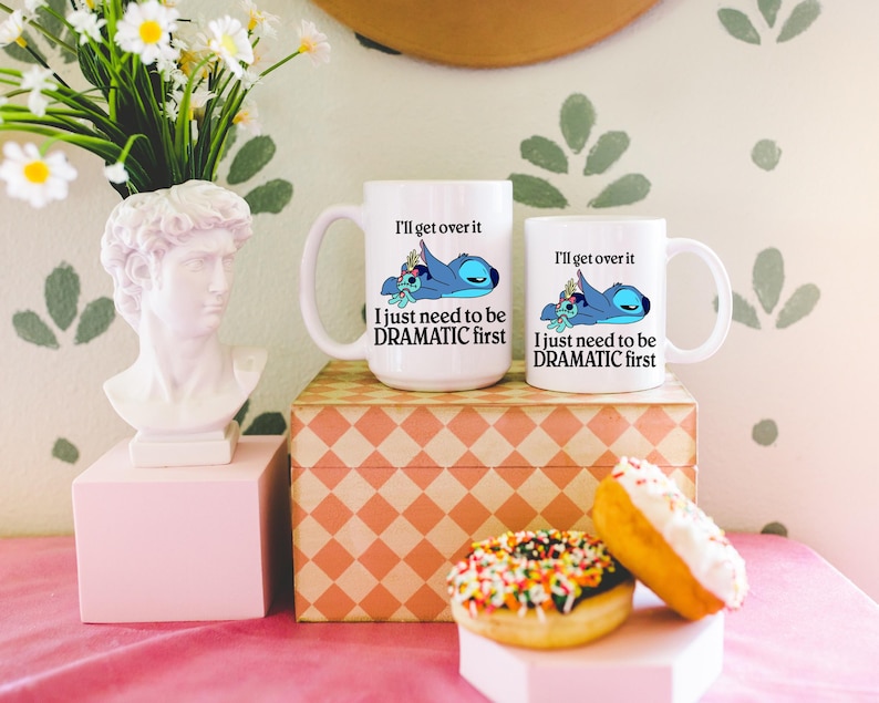 two white mugs with a cartoon character and text, placed on a pink tablecloth. There are also two doughnuts with colorful sprinkles and a white bust of a person on a pedestal. The background has a floral pattern.