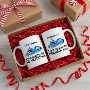 A pair of white mugs with a humorous message printed on them, placed in a brown gift box filled with red shredded paper and red ribbons.