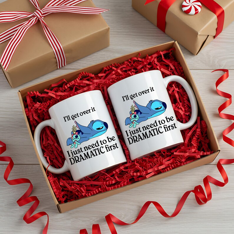 A pair of white mugs with a humorous message printed on them, placed in a brown gift box filled with red shredded paper and red ribbons.