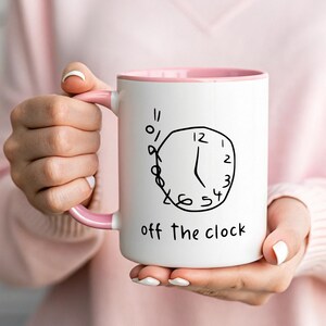 May include: White ceramic mug with a pink interior and handle. The mug features a hand-drawn clock design in black ink, with the words "off the clock" below the clock face. The mug is held by a person.