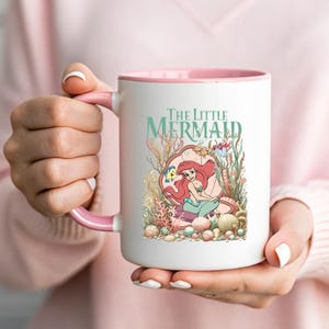 A person is holding a white mug with a pink interior and a picture of a mermaid on it.