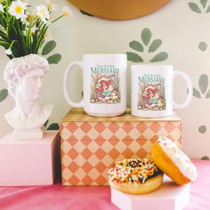 a pink table with a bust of a man, two mugs with a mermaid design, a box, a vase with flowers, and two donuts with colorful sprinkles.