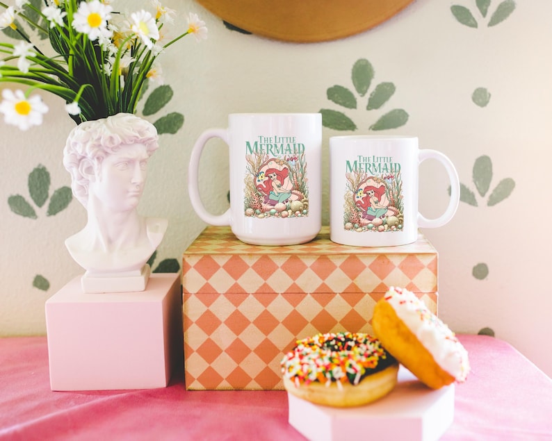 a pink table with a bust of a man, two mugs with a mermaid design, a box, a vase with flowers, and two donuts with colorful sprinkles.