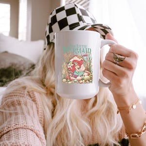 A person holding a mug with a mermaid design, wearing a checkered cap and a pink sweater.