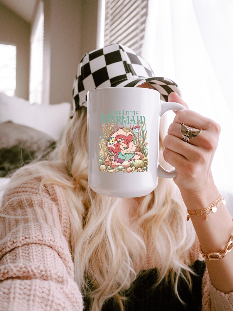 A person holding a mug with a mermaid design, wearing a checkered cap and a pink sweater.