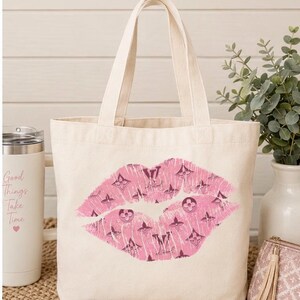 K&ouml;nnte beinhalten: Eine cremefarbene Canvas-Tragetasche mit einer gro&szlig;en, rosafarbenen Grafik von Lippen im Used-Look. Das Lippendesign ist mit einem sich wiederholenden Muster aus dunkelbraunen floralen und geometrischen Symbolen gef&uuml;llt, darunter LV-Buchstaben.