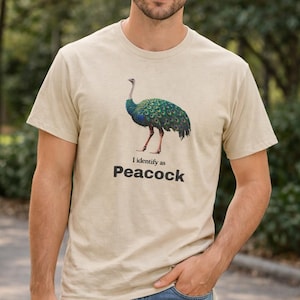 May include: Beige t-shirt with a peacock illustration in green, blue, and brown. The text "I identify as Peacock" is printed below the image. The shirt is short-sleeved and made of a soft material.