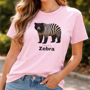 May include: A light pink t-shirt featuring a graphic of a bear with zebra stripes. The text "I identify as Zebra" is printed below the image in black. The shirt is made of a soft material.
