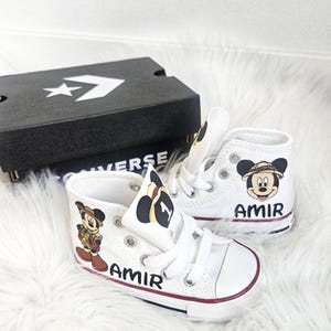 May include: White high-top sneakers with Mickey Mouse graphics and the name "AMIR" printed on the side. The shoes have white laces, a red stripe on the sole, and are next to a black Converse box.