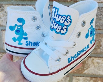 May include: White high-top canvas shoes with blue and white "Blue's Clues" logo and a blue cartoon dog on each shoe. The shoes are personalized with the name "Sherman" on one shoe and "Sher" on the other.