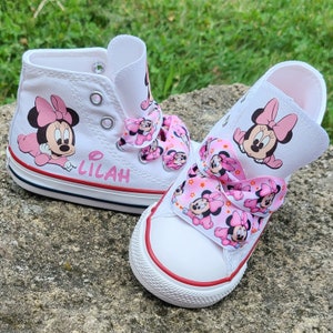 May include: A pair of white canvas shoes with pink Minnie Mouse designs. The shoes have pink ribbon laces and the name "Lilah" written on one shoe.