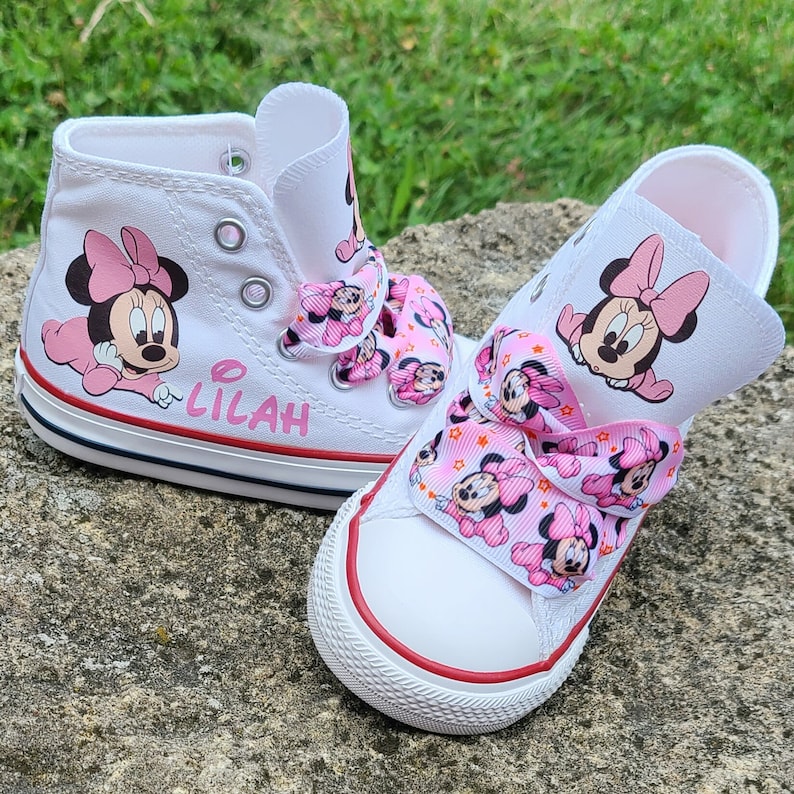 May include: A pair of white canvas shoes with pink Minnie Mouse designs. The shoes have pink ribbon laces and the name "Lilah" written on one shoe.