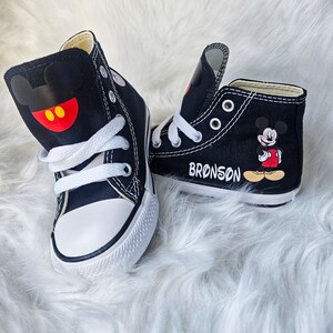 May include: Pair of black high-top sneakers with white laces and soles. One shoe features a Mickey Mouse design on the side and the other has a Mickey Mouse head design. The name "BRONSON" is printed on one shoe.