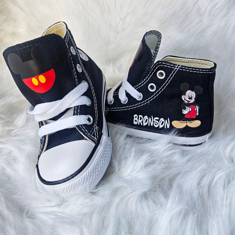May include: Pair of black high-top sneakers with white laces and soles. One shoe features a Mickey Mouse design on the side and the other has a Mickey Mouse head design. The name "BRONSON" is printed on one shoe.