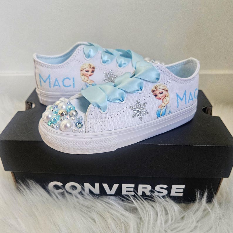 May include: White canvas sneakers featuring light blue ribbon laces, a cartoon character illustration, silver snowflake accents, and the name Maci in blue text. The toe caps are decorated with an assortment of white, blue, and silver pearls.